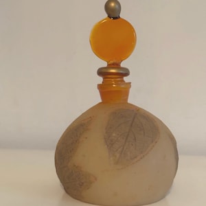 Puede incluir: Un frasco de perfume vintage con un cuerpo redondeado esmerilado con diseños de hojas. El frasco tiene un tapón transparente de color naranja con una parte superior circular y una pequeña esfera dorada. El frasco es probablemente de vidrio.