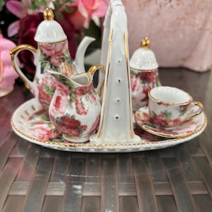 Könnte beinhalten: Ein Miniatur-Porzellan-Tee-Set mit floralem Design, bestehend aus Teekanne, Milchkännchen, Zuckerdose, Teetasse und Untertasse. Das Set befindet sich auf einem weißen Tablett mit goldenem Griff.