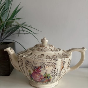 Könnte beinhalten: Eine cremefarbene Teekanne mit einem goldenen Blumenmuster und einer Szene mit einer Frau in einem rosa Kleid. Die Teekanne hat einen runden Körper und einen Deckel mit einem Knopf.