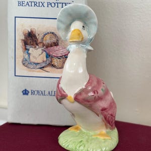 May include: A ceramic goose figurine wearing a blue bonnet and a pink dress, standing on a patch of green grass. The figurine is from the "World of Beatrix Potter" collection by Royal Albert.