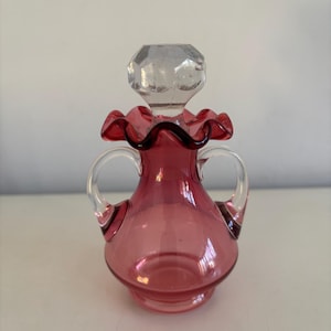 Puede incluir: Una pequeña botella de vidrio rojo rubí con un tapón transparente facetado. La botella tiene un cuello con volantes y dos asas claras y arqueadas. La base es de un tono de rojo ligeramente más oscuro. Una pieza decorativa.
