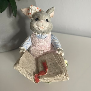 Può includere: Una statuetta di coniglio grigia che indossa un vestito rosa e una fascia bianca con pois rossi, seduta su un libro aperto con un segnalibro a nastro rosso.