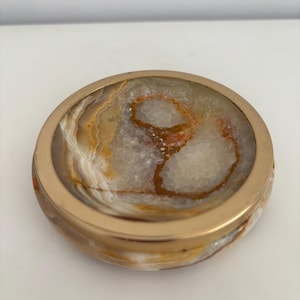 Könnte beinhalten: Ein runder, dekorativer Aschenbecher mit einem goldfarbenen Metallrand. Der Innenraum weist ein Natursteinmuster in Weiß-, Braun- und Orangetönen auf. Der Aschenbecher ist ein stilvolles und funktionales Element für die Inneneinrichtung.