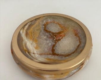 Puede incluir: Un cenicero redondo decorativo con un borde de metal dorado. El interior presenta un patrón de piedra natural con tonos de blanco, marrón y naranja. Este cenicero es un artículo elegante y funcional para la decoración del hogar.