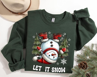 May include: Dark green sweatshirt featuring a whimsical snowman design. The snowman is wearing a red hat, mittens, and boots, with a white scarf. The text "LET IT SNOW" is printed in white. Christmas-themed apparel.
