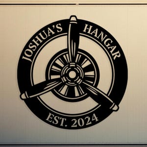 May include: Black circular sign with a propeller design and the text "JOSHUA'S HANGAR EST. 2024". The sign is mounted on a white hangar door. A small airplane is visible in the background.