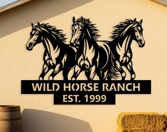 Può includere: Cartello in metallo nero raffigurante tre cavalli in corsa, con la scritta "WILD HORSE RANCH EST. 1999". Il cartello è montato su un esterno di edificio beige, con una botte e balle di fieno in primo piano.
