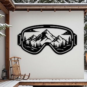 May include: Black metal wall art showing ski goggles with a mountain and forest scene. The artwork is mounted on a white wall outside a house with snow-covered steps and a wooden deck. A sled and skis are nearby.