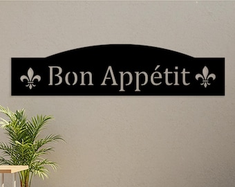 Puede incluir: Letrero de metal negro con las palabras "Bon Appétit" en blanco, flanqueado por símbolos de flor de lis. El letrero está montado en una pared de color claro, con una planta y asientos al aire libre en el fondo.
