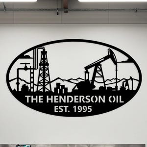 May include: Black metal wall art featuring an oil field silhouette within an oval frame. The design includes oil derricks, pipelines, and the text "THE HENDERSON OIL EST. 1995". The artwork is mounted on a white wall.