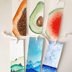 Puede incluir: Seis marcapáginas rectangulares de papel con borlas de cuerda blanca. La fila superior presenta ilustraciones en acuarela de una rodaja de papaya, medio aguacate y una rodaja de sandía. La fila inferior muestra colinas verdes, montañas bajo un cielo nocturno y una playa.