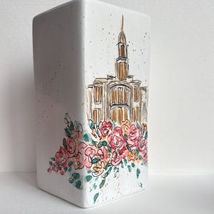 May include: A white rectangular vase featuring a hand-painted illustration of a building with a tall spire and gold detailing. The vase is adorned with a floral arrangement of pink and peach roses and green foliage.
