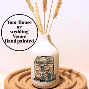 May include: A white, hand-painted vase featuring a detailed illustration of a stone house with a green door and a gray roof. The vase holds dried wheat stalks. Text on the image reads "Your House or wedding Venue Hand painted". The vase sits on a cork base.