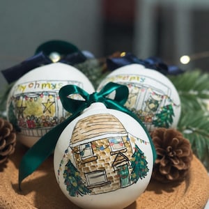 May include: Three white spherical ornaments featuring hand-painted illustrations of houses decorated for the holidays. Each ornament is topped with a dark green ribbon bow and displays a detailed scene of a brick home with string lights and trees.