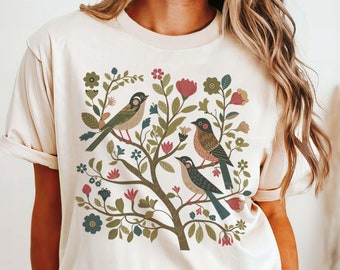 May include: A cream colored t-shirt with a floral design featuring three birds and a tree. The design is in shades of green, blue, red, and brown.