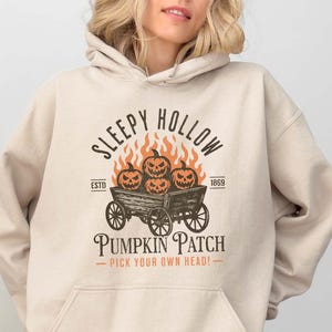 May include: Sand-colored hoodie with a front graphic featuring the text "Sleepy Hollow Pumpkin Patch" and a design of pumpkins in a wagon with flames. The hoodie has a hood and a front pocket.