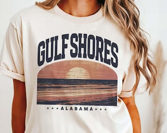 May include: Ivory-colored t-shirt featuring a sunset graphic with the words "GULF SHORES" above "ALABAMA". The graphic depicts a sun setting over the ocean with shades of orange, pink, and blue. The shirt is a relaxed fit.