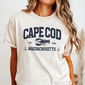 May include: A cream-colored t-shirt with a navy blue graphic that says "CAPE COD" with a lobster and anchor design. The text below the graphic says "EST. 1602 MASSACHUSETTS".