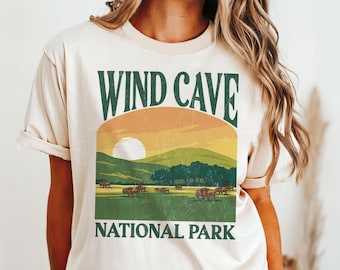 May include: A beige t-shirt with a vintage-style graphic of Wind Cave National Park. The graphic features a sunset over a mountain range with bison in the foreground. The text "Wind Cave National Park" is printed in green.