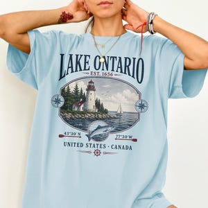 May include: A light blue t-shirt featuring a graphic of a lighthouse on a rocky shore, a sailboat, and a fish. The text reads LAKE ONTARIO EST. 1656, 43°30'N 77°30'W, and UNITED STATES CANADA. The design includes compass rose icons and oars.
