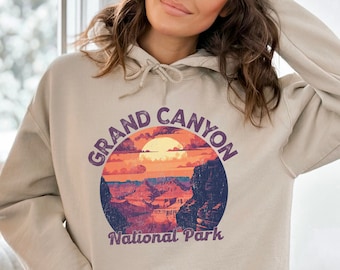 May include: Sand-colored hoodie featuring a graphic of the Grand Canyon National Park. The design includes a sunset scene with the text "GRAND CANYON" and "National Park" in a circular layout. The hoodie has a front pocket and a drawstring hood.