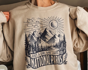 Könnte beinhalten: Ein beiger Pullover mit einem grafischen Aufdruck von Bergen, einer Sonne und Wolken. Der Text "Just a girl who loves National Parks" ist auf dem Pullover aufgedruckt.
