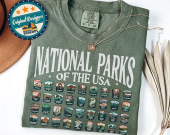 May include: A green t-shirt with a white graphic of all 63 National Parks of the USA. The graphic includes the name of each park and a small illustration of the park's logo. The text "All 63 National Park Badges" is printed below the graphic.