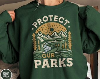 May include: Forest green sweatshirt with the text "PROTECT OUR PARKS" in a vintage-style design. The graphic features a mountain range, river, and trees under a sunburst. The sweatshirt is a solid color.