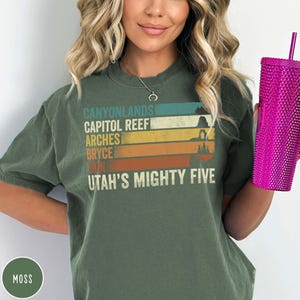 May include: Moss green t-shirt with the text "Canyonlands, Capitol Reef, Arches, Bryce, Zion, Utah's Mighty Five" in a retro design. The shirt has a crew neck and short sleeves. A pink tumbler with a straw is held in the hand.
