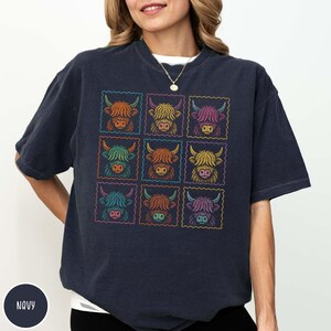 May include: Navy blue t-shirt featuring a grid of nine colorful Highland cow illustrations. Each cow is framed within a square, with vibrant rainbow-colored outlines. The shirt has a relaxed fit and a crew neck.
