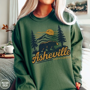 May include: A military green sweatshirt with a graphic of a bear, mountains, and a sun. The word "Asheville" is in a retro font, with "North Carolina" below. The sweatshirt is a solid colour.