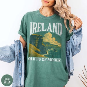 May include: Light green t-shirt with the word "IRELAND" above a graphic of the Cliffs of Moher. The text "CLIFFS OF MOHER" is below the graphic. The shirt has rolled-up sleeves and is paired with a light blue denim jacket.