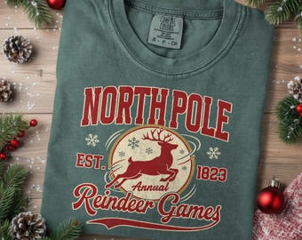 May include: A folded, green t-shirt with a red graphic that reads "North Pole Annual Reindeer Games." The design includes a reindeer, snowflakes, and the year 1823. The shirt is on a wooden surface with Christmas decorations.
