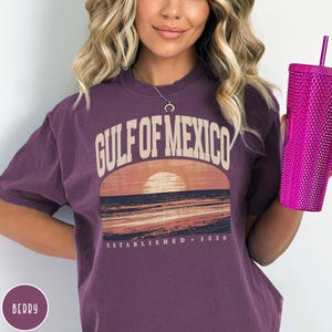 May include: A purple t-shirt with a vintage-style graphic of a sunset over the Gulf of Mexico. The text "Gulf of Mexico" is in a retro font and the text "Established 1550" is below the graphic.