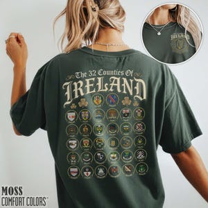 May include: Dark green t-shirt with the text "The 32 Counties Of Ireland" and a detailed graphic of county emblems. The shirt also features the text "MOSS COMFORT COLORS" at the bottom. A smaller graphic of the Ireland emblem is on the upper right.