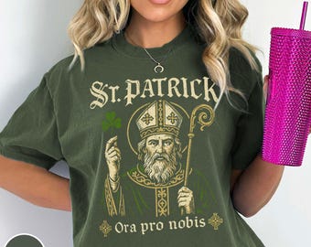 Puede incluir: Camiseta verde oliva del Día de San Patricio con un gráfico de San Patricio sosteniendo un trébol y un báculo, con el texto "St. Patrick" y "Ora pro nobis" en letras doradas. Se sostiene un vaso rosa con pajita.