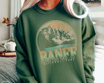 May include: A military green sweatshirt featuring a circular graphic of mountains and the text "BANFF NATIONAL PARK" in a retro font. The sweatshirt has long sleeves and a crew neck. The words "MILITARY GREEN" are printed on the bottom left.