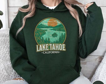 May include: Forest green hoodie featuring a circular graphic of Lake Tahoe with a sunset, trees, and water. The text "LAKE TAHOE CALIFORNIA" is printed below the graphic in a vintage style. The hoodie has a front pocket and a hood.