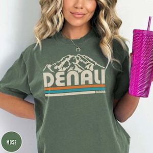 May include: A moss green t-shirt with a vintage-style graphic of a mountain range and the text "DENALI ALASKA".