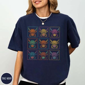 May include: Navy blue t-shirt featuring a grid of nine colorful Highland cow illustrations. Each cow is framed within a postage stamp-like border. The shirt has a relaxed fit and short sleeves. The text "TRUE NAVY" is printed on the bottom left.