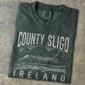 May include: Dark green t-shirt with the text "COUNTY SLIGO IRELAND" in white. The shirt features a graphic of a mountain range and clouds. The shirt is folded on a textured surface.