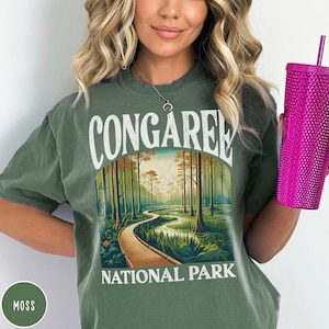 May include: A green t-shirt with a vintage-style graphic of Congaree National Park. The graphic features a boardwalk winding through a swampy forest with tall trees and a blue sky.