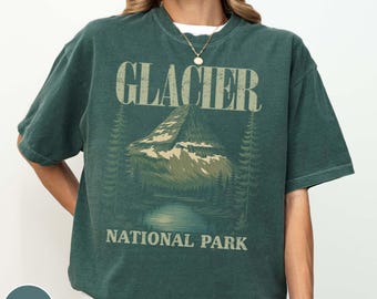 May include: A dark green t-shirt with the words "GLACIER NATIONAL PARK" printed on the front. The graphic features a mountain scene with trees and a lake. The shirt has a relaxed fit.