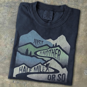 May include: Dark navy blue t-shirt with a mountain graphic and the text "Just Another Half Mile or So." The design features a hiker on a trail, trees, and layered mountains in shades of blue and green.