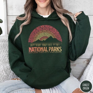 May include: A dark green hooded sweatshirt with a vintage-style graphic of mountains and trees. The graphic features the text "National Parks" and "Explore Protect Preserve". The graphic also includes the names of many national parks in the United States.
