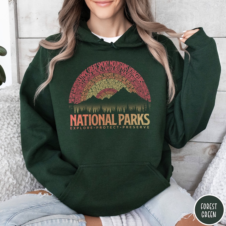 May include: A dark green hooded sweatshirt with a vintage-style graphic of mountains and trees. The graphic features the text "National Parks" and "Explore Protect Preserve". The graphic also includes the names of many national parks in the United States.