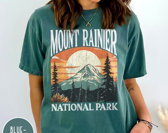 May include: A green t-shirt with a vintage-style graphic of Mount Rainier National Park. The graphic features a mountain, a sunset, and trees. The text "Mount Rainier National Park" is printed in white.