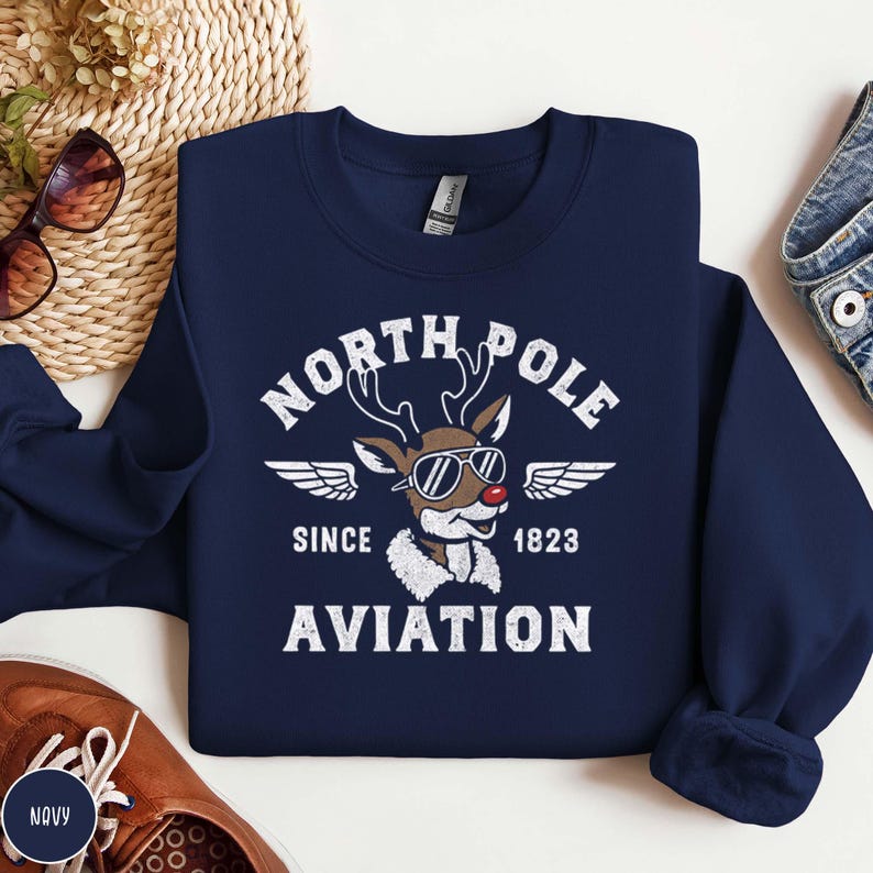 May include: Navy blue sweatshirt with a white graphic featuring a reindeer wearing sunglasses and the text "NORTH POLE AVIATION SINCE 1823". The reindeer has a red nose and is wearing a white scarf.