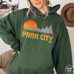May include: A green hooded sweatshirt with a retro-style graphic design featuring a sunset, mountains, and the text "Park City, Utah".