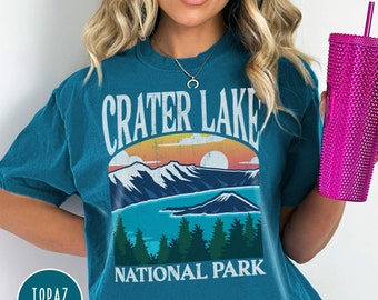 May include: A turquoise blue t-shirt with a vintage-style graphic of Crater Lake National Park. The graphic features a sunset over a mountain range with a lake in the foreground. The text "Crater Lake National Park" is printed in white below the graphic.